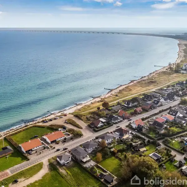 Hav spiller stille ved kystlinjen, mens en bro strækker sig i horisonten. Forstadsbygninger ligger langs veje parallelt med sandstranden, og en sportshal ses til højre.