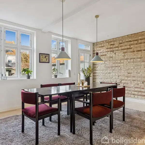 Spisebord med fire stole står på et tæppe. Hængende lamper over bordet. Rummet har en murstensvæg og store vinduer med udsigt til en gårdhave. Plantet i hjørnerne.