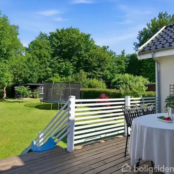 Rundt bord med dug og blomster står på en terrasse med stole. I baggrunden ses en stor, grøn have med træer og en trampolin under en blå himmel.