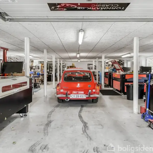 En rød bil med nummerplade CY 28 957 står parkeret i en rummelig garage fyldt med værktøj, hylder og udstyr, hvilket indikerer et værkstedmiljø.