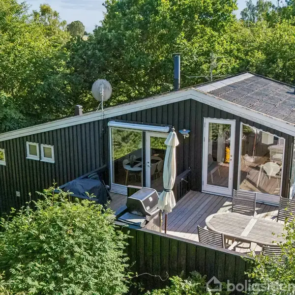 Et sort træhus har en terrasse med en grill og rundt bord. Det ligger omgivet af frodig grøn vegetation. Sollys skinner gennem glasdørene, hvilket giver et venligt indtryk.