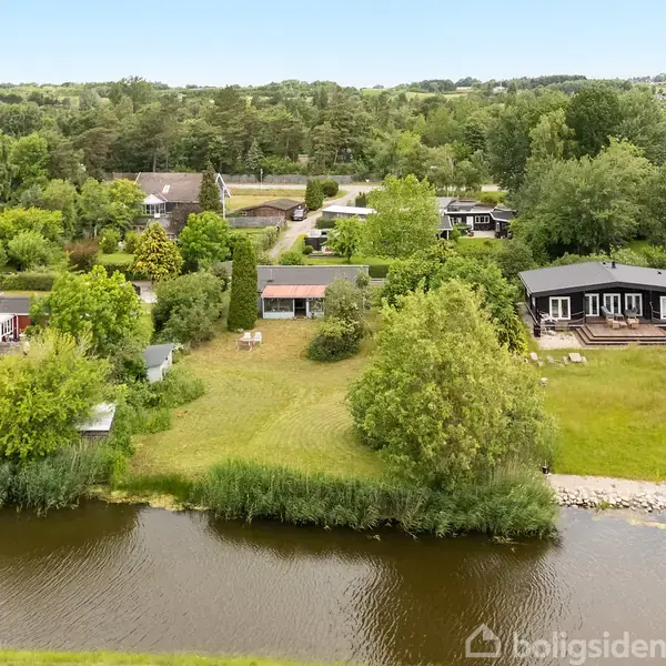 Huse ligger ved en flod, omgivet af grønne haver med træer og buske. Hovedhuset har en terrasse og udsigt til vandet, omgivet af en frodig, skovrig baggrund.