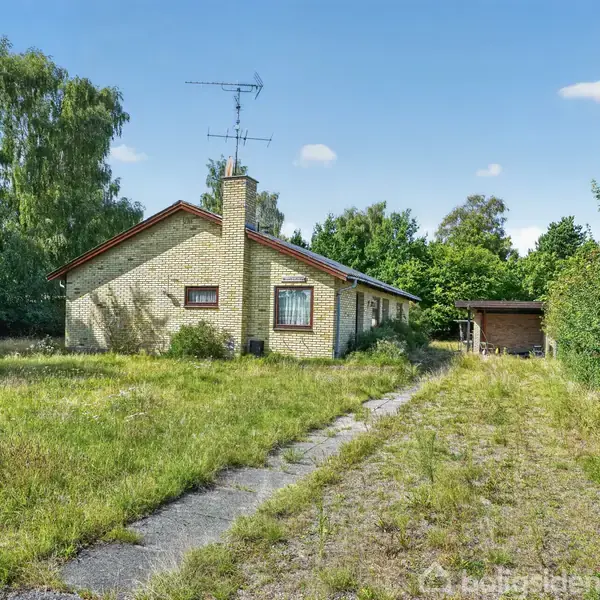 Huset står i en have med høj, vildtvoksende græs. En smal sti fører til en garage i baggrunden. Træer og buske omgiver området under en klar, blå himmel.