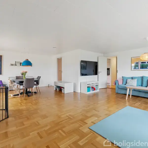 Stue med turkis sofa, der står ved et sofabord på et lyseblåt tæppe. Tv er monteret på væggen. Bagved ses spisebord med stole under en blå lampe. Trægulv omgiver.