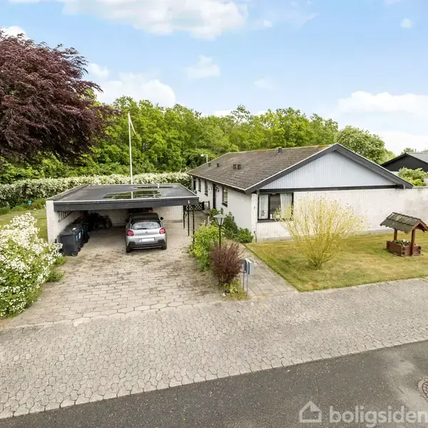 Bil holder parkeret under en carport ved siden af et hvidt hus med have og flagstang; omgivet af træer og buske på en rolig villavej.
