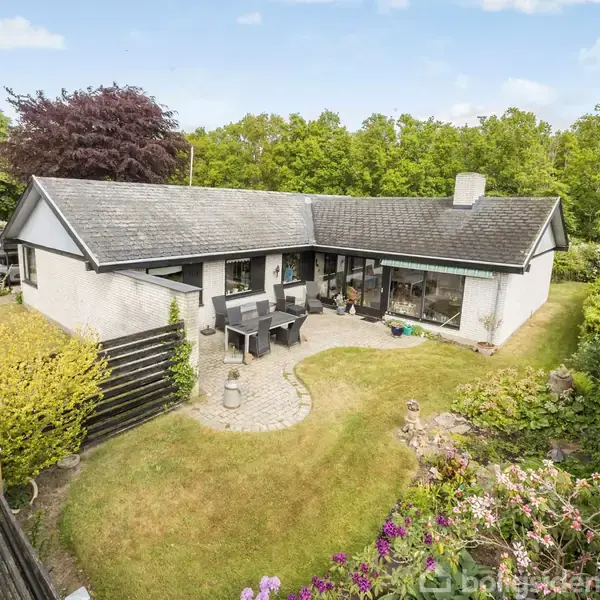 Huset er et enkelt-etagers, hvidt murstenshus med mørkt tag, beliggende i en frodig have med havesæt på terrasse. Omkranset af træer og buske, skaber en rolig, grøn atmosfære.