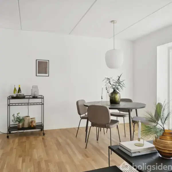 Et spisebord med stole står i et lyst rum med trægulv. En barvogn med flasker og planter er langs væggen, og et stort vindue giver udsigt.