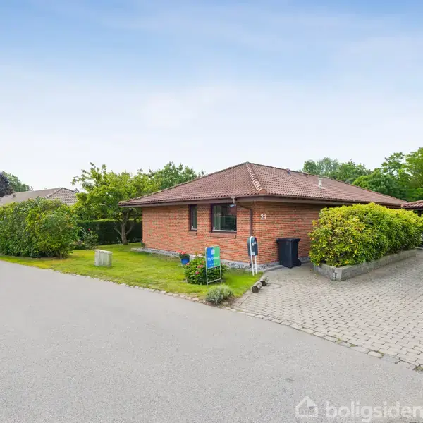 Et rødt murstenshus med carport står til venstre for en vej. Omgivet af en trimmet græsplæne og buske, ses flere huse og grønne træer i baggrunden.