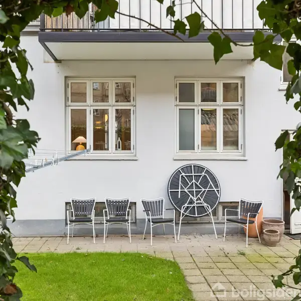 Hvide kurvestole står opstillet langs en hvid husmur med to vinduer, omkranset af grønne blade fra en have, der skaber en hyggelig og indbydende atmosfære.