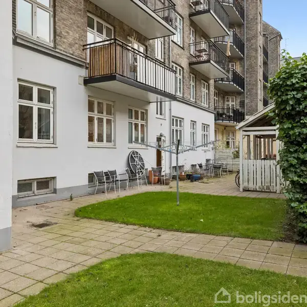 Baggård med græsplæne, flisebelagt sti og cykel ved et træ. Omkranset af et flerfamiliehus med balkoner, havebord og stole ved siden af en lille pavillon.