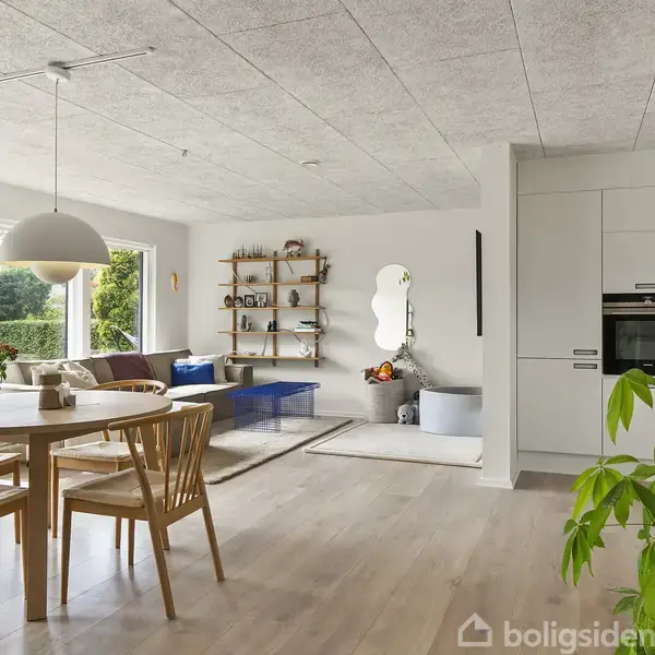 Et lyst, åbent rum med spisestue og stue. Rundt spisebord med stole, sofa med sofabord. Reol og spejl på væggen, store vinduer mod haven. Ingen tekst.