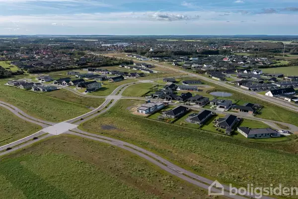 Luftfoto viser et boligområde med moderne huse, spredt langs snoede veje gennem et grønt landskab. Området ligger i nærheden af en skov og andre bygninger.