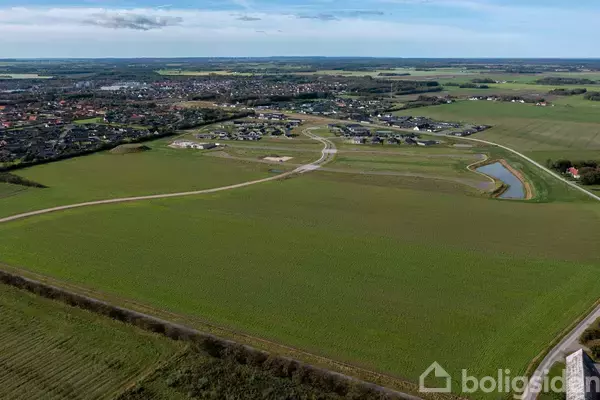 Mark med grønne marker omkranser en vej, der snor sig mod en nærliggende by. En lille sø ligger i højre hjørne, og himlen er overskyet.