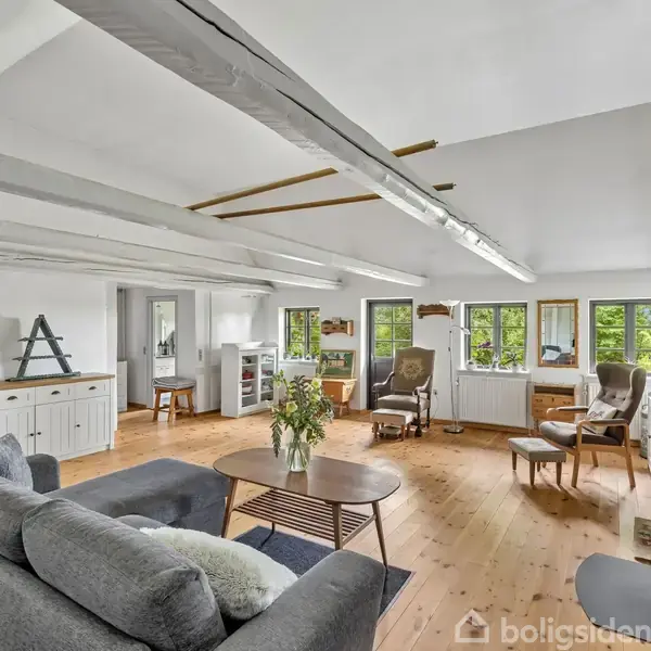 Stue med grå sofa, rundt sofabord og blomstervase, omgivet af lyse træmøbler og vinduer med udsigt til skov. Højt til loftet med synlige bjælker og trægulv.