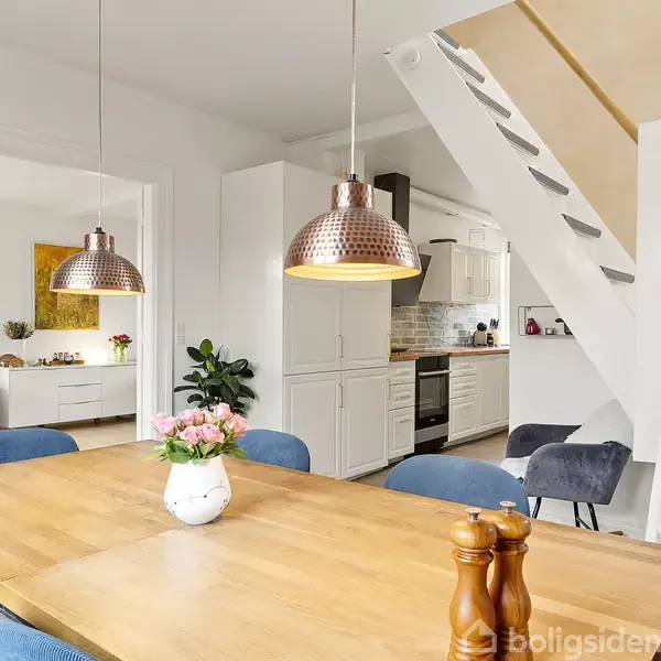 Spisebord med blomster i vase og peberkværn står under kobberlamper. Ved siden er trappe op, og i baggrunden ses et lyst køkken med komfur og skabe.