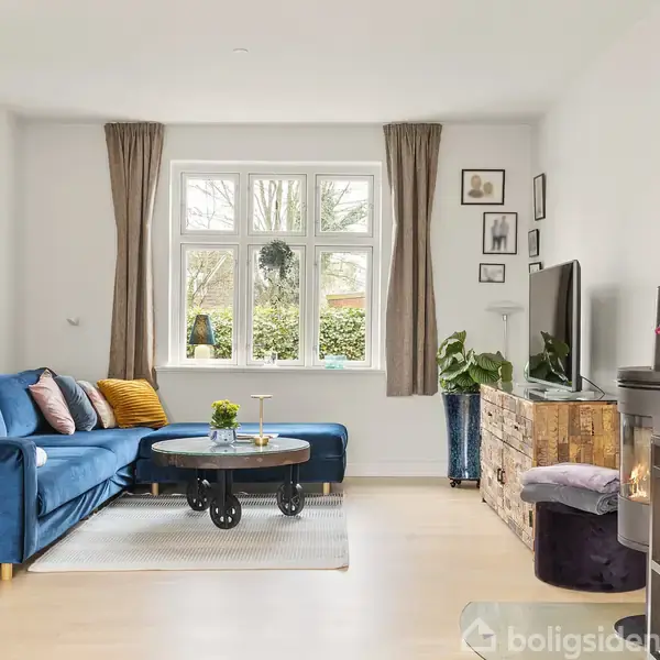 Blå sofa med farverige puder og sofabord står foran et stort vindue med gardiner. Brændeovn, reol, planter og billeder danner en hyggelig stueindretning. Ingen tekst fundet.