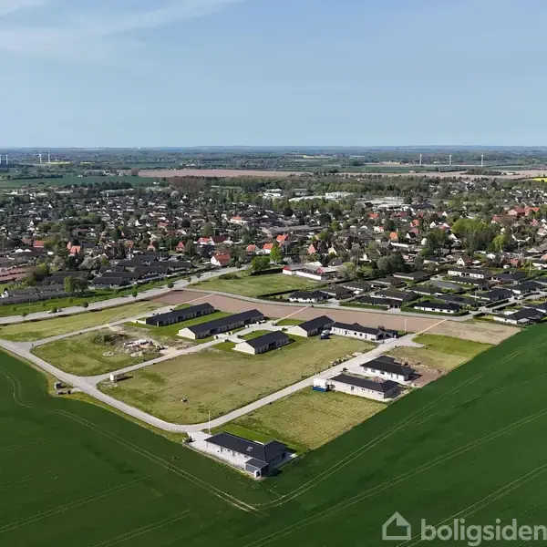 Nyopførte huse står stille på en åben grund ved kanten af en by. Omkringliggende marker og et vidstrakt syn af byens huse og veje strækker sig til horisonten.
