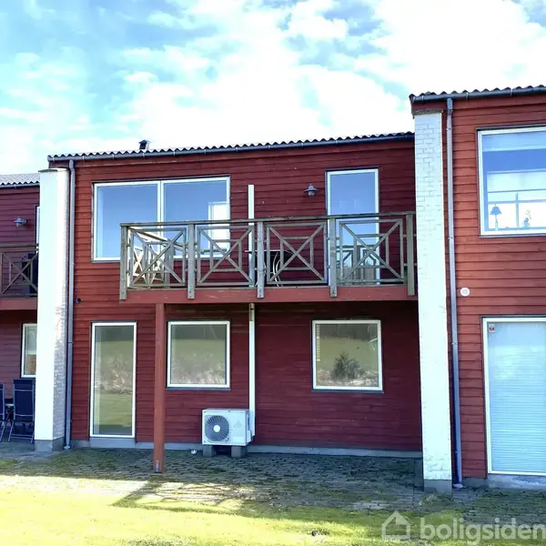 Rødt træhus med store vinduer og altaner. Huset står i en græsbeklædt have under en blå himmel. En airconditionenhed er monteret på facaden.