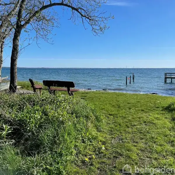 Bænk står stille ved kysten foran en træbro, omgivet af græs og træer. Havet strækker sig ud i horisonten under en klar blå himmel.