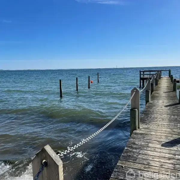 En træmole strækker sig ud over havet, omgivet af blidt bølgende vand. Klar blå himmel ovenover i dagslys. Flere pæle står i vandet ved siden af molen.
