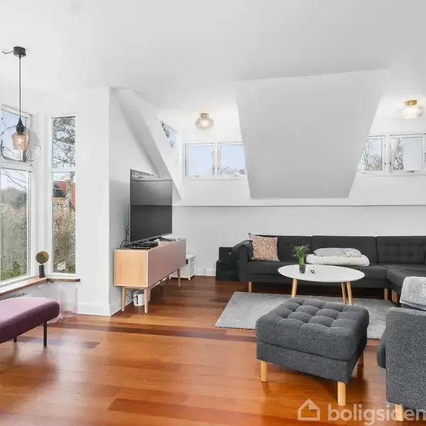 Stue med mørkegrå sofa i hjørnet, sofabord og lille skammel. Store vinduer lukker lys ind, træplankegulv. Dekorationer omfatter puder, planter, en lampe og lysestager.