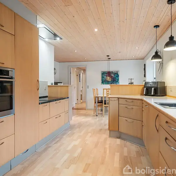 Køkken med lyse træskabe og indbyggede apparater, inklusiv ovn og komfur. Rummet er lyst med træloft og lyse gulve. Belysningen er moderne, og der er udsigt til en spisestue.