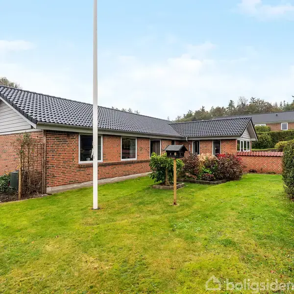 Et rødt murstenshus med sort tag står i en velplejet have. Græsplænen er omgivet af buske, og en flagstang står i forgrunden. Skoven ses i baggrunden.