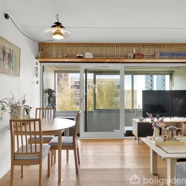 Et spisebord med stole står i en lys stue. Et sofabord og en sofa er placeret mod en skydedør, der åbner ud til en balkon med udsigt.