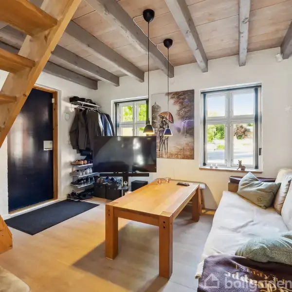 En stue med en trætrappe til venstre, en sofa og sofabord i midten, en fladskærms-TV foran vinduerne, omgivet af moderne dekorationer og lyse trægulve.