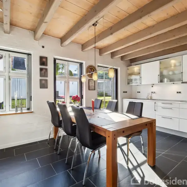Træbord med sorte stole står i et lyst køkken med hvide skabe og sort flisegulv. Sollys strømmer ind gennem vinduerne, fremhæver de synlige loftsbjælker og skaber en indbydende atmosfære.