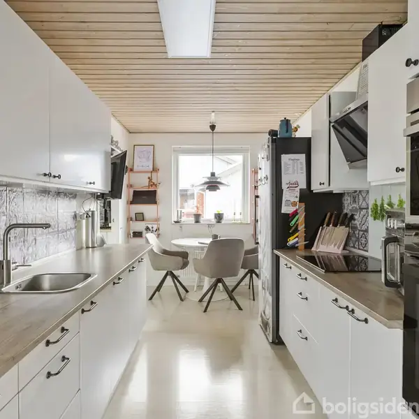 Moderne køkken med hvide skabe langs begge sider. Indbygget ovn og kogeplade til højre. Centreret spiseområde med bord og fire stole ved et vindue. Dekorativ mosaik-vægflise.