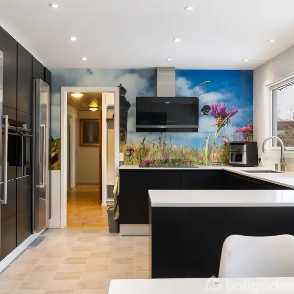 Et moderne køkken med sorte skabe og en stor køleskab. En farverig vægdekoration med blomster bagved vasken. Store vinduer giver naturligt lys. Ingen synlig tekst.