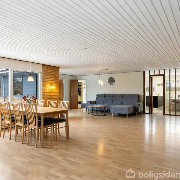 Spisestuebord med stole står i et lyst, åbent rum med trægulv og hvidt loft. I baggrunden er en sofa og store vinduer, der giver udsigt til en terrasse.