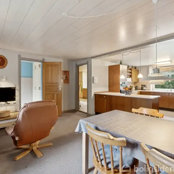 To lænestole står foran et tv i en hyggelig stue, der er forbundet med et åbent køkkenområde. Spisebordet står nær køkkenet, der har brune skabe.