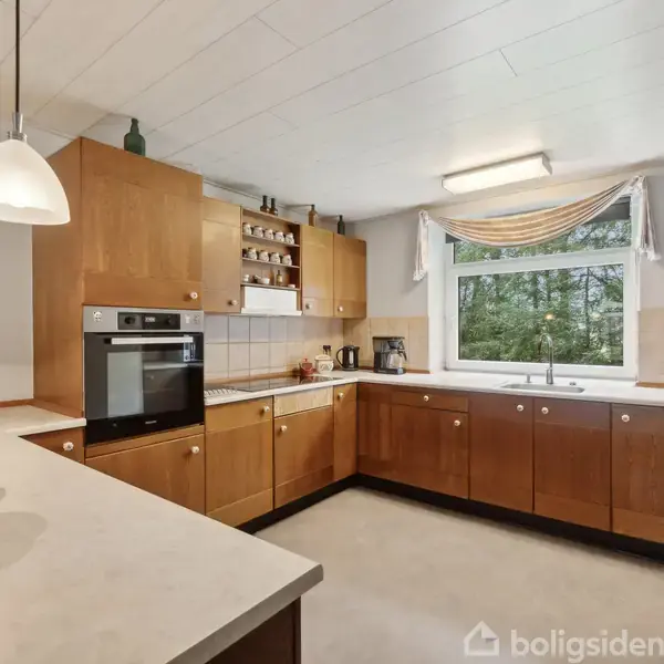 En moderne køkkenindretning med træskabe og indbygget ovn. Der står køkkenudstyr på køkkenbordet. Et stort vindue med gardiner giver udsigt til grønne træer.