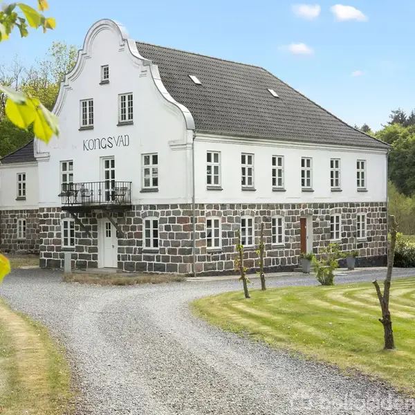 En hvid, gammel bygning med sort tegltag, skiltet “Kongsvad” ovenfor en balkon, står i en grøn, velplejet have med en grusvej forrest og skov i baggrunden.