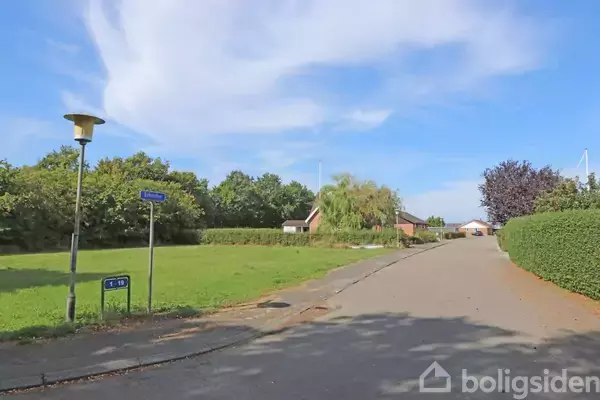 Gadelygte står ved vejskilt med teksten "Rolighedsvej". Vejen strækker sig gennem et grønt kvarter med hække, træer og enkelte bygninger i baggrunden under en blå himmel.