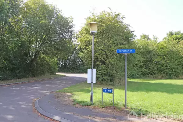 En gadelampe står ved siden af et vejskilt med teksten "Birketoften" og et andet skilt med "1-19" nær en vejkurve. Omgivet af grønne buske og træer.