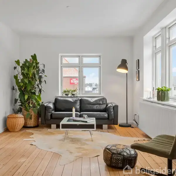 En sort sofa står foran et vindue, flankeret af en stor plante og en gulvlampe. Et tæppe ligger på trægulvet, mens en grøn stol og et lille bord supplerer rummet.
