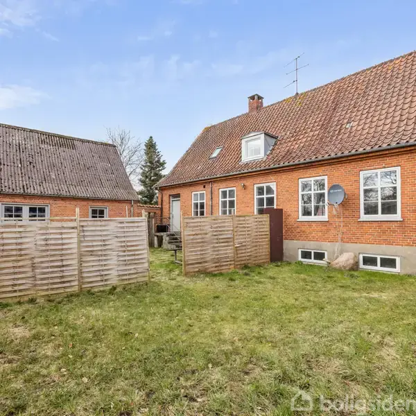 Rødt murstenshus med skråt tag, omgivet af græsplæne og træhegn. Nabobygning til venstre med lignende stil. Blå himmel i baggrunden.