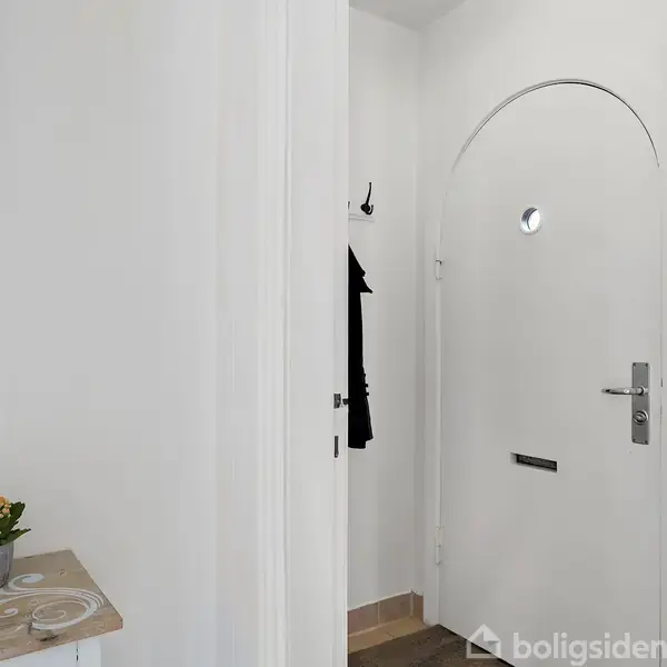 Dør med rundt vindue står lukket i en hvid entré med træbænk og en lille plante. På væggen hænger skiltet "HOME is where we share it with" og en sort jakke.