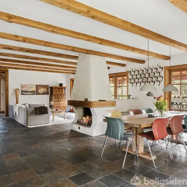 Moderne stue med åben pejs centralt placeret, flammerne brænder livligt. Et spisebord omgivet af farverige stole. Trægulve og loftsbjælker tilføjer en rustik charme, med udsigt til balkon.