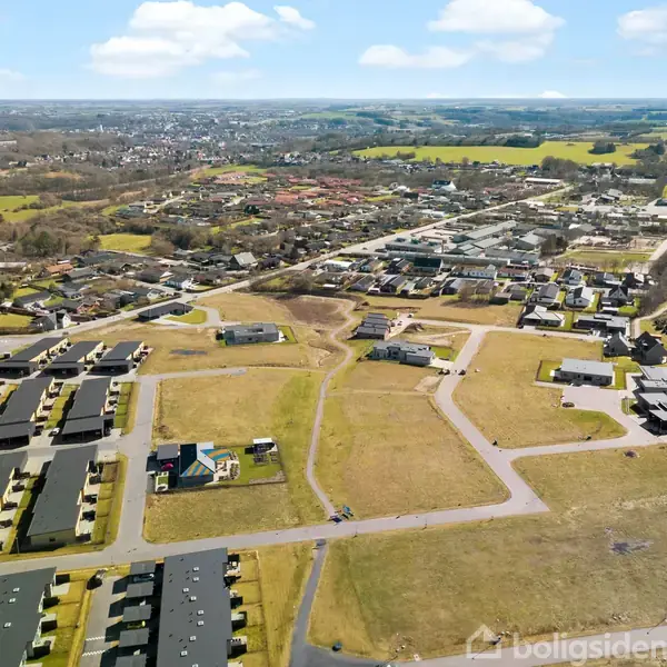 Luftfoto viser et boligområde med rækkehuse og veje, omgivet af åbne marker og en by i baggrunden under en klar himmel.