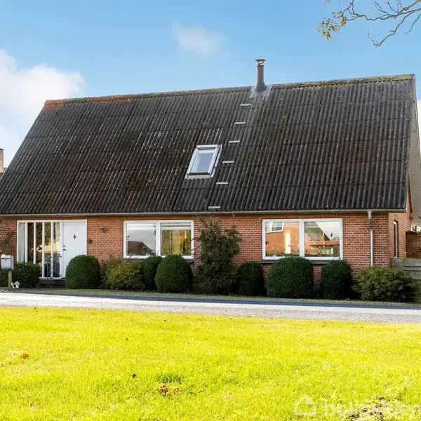 Rødstenshus står stille omgivet af en velplejet græsplæne og buske under en blå himmel. Huset har et skråt sort tag med flere vinduer placeret i en linje.