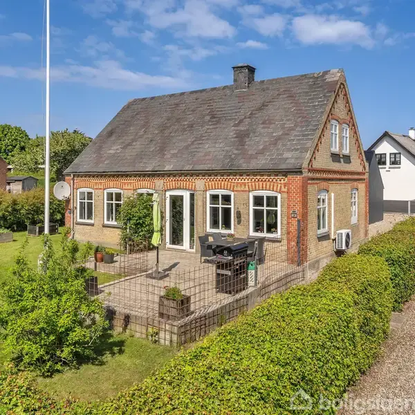 Et rødt murstenshus står omgivet af en grøn have med hæk. Huset har sorte tagsten, hvide vinduesrammer, og en terrasse med havemøbler. Det er en solrig dag med blå himmel.