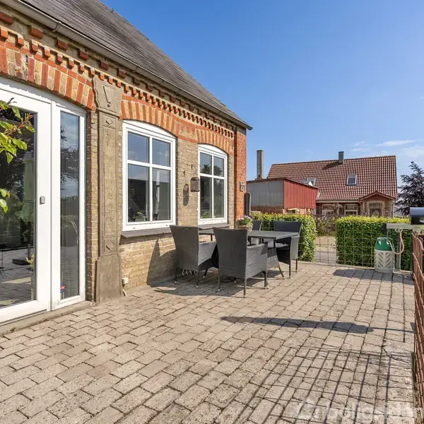 Murstenshus med terrasse. Et bord og fire stole står til højre for bagdør og vinduer. Parasol folder sammen ved husets væg. Grønt hegn og nabolag i baggrunden.