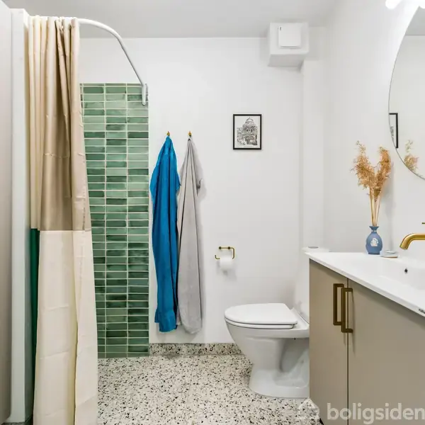 Badeværelse med bruser, grøn flisevæg og bruseforhæng. Ved siden af står et toilet med håndklæder på kroge. En vask med spejl og sæbedispenser findes til højre. Enkelt dekoreret.