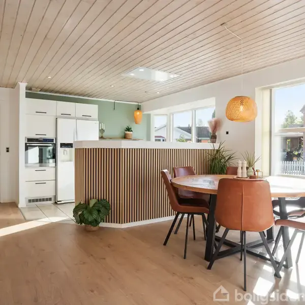Spisebord med læderstole står ved vindue, omgivet af en lys moderne køkkenindretning med træloft og planter. Sollys strømmer ind gennem store vinduer, skaber en indbydende atmosfære.