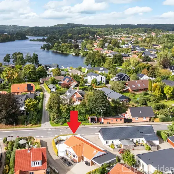 En række mindre huse ses fra fugleperspektiv, mens en rød pil peger på et specifikt hus. Omgivelserne inkluderer veje, grønne områder og en stor sø.