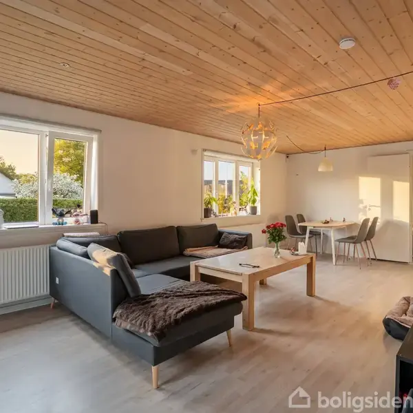 Sofaen er placeret i en stue, med et sofabord foran og en spisebordgruppe i baggrunden. Værelset er lyst med trægulv og loft.